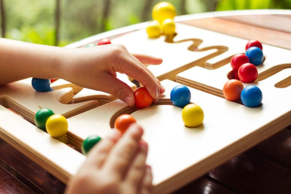 Child's hands maneuver colorful wooden balls through a maze on a wooden puzzle board.