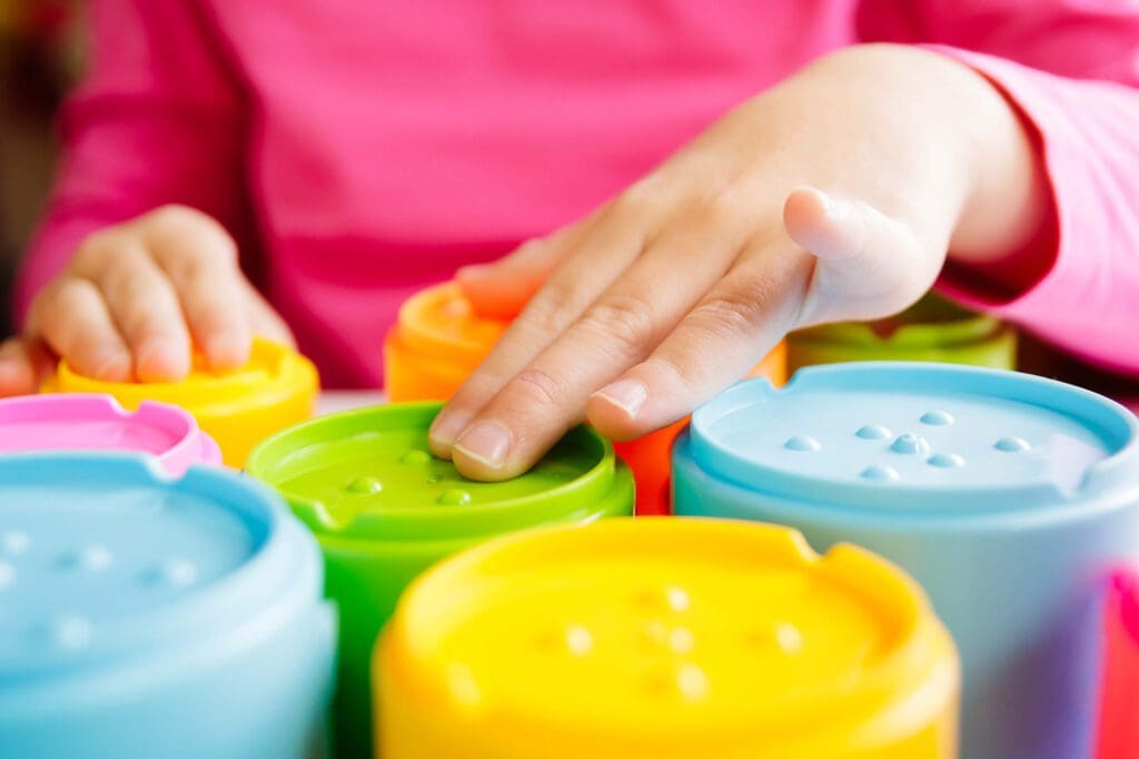 A child's hand touching a colorful set of tactile plastic containers with embossed dots, including blue, green, and yellow lids.