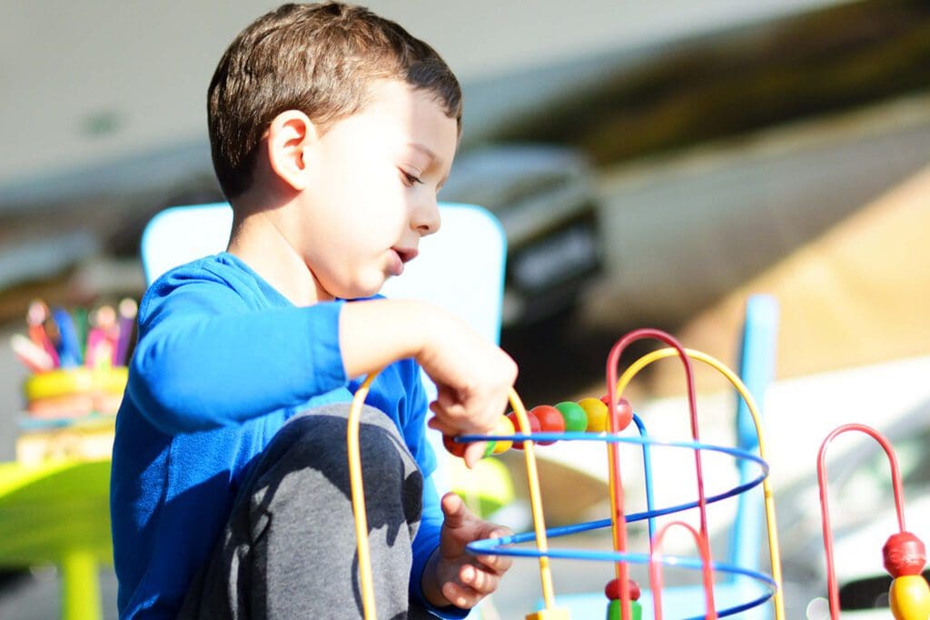 Child in blue shirt sitting and playing with a colorful wire bead maze toy.