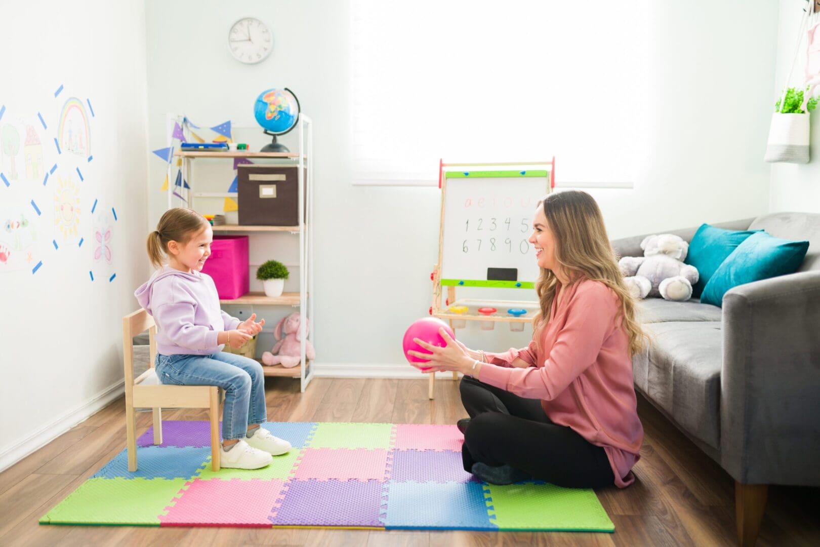 A woman and a young girl playing with a pink ball in a colorful, well-lit room with educational toys and a globe.