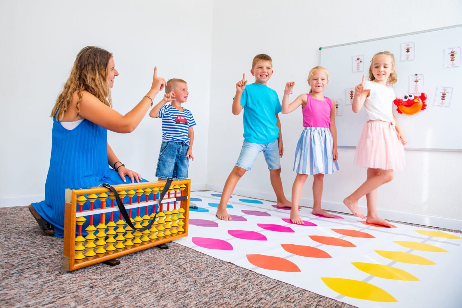Children playing a game on a colorful Twister mat while a woman watches and cheers them on.