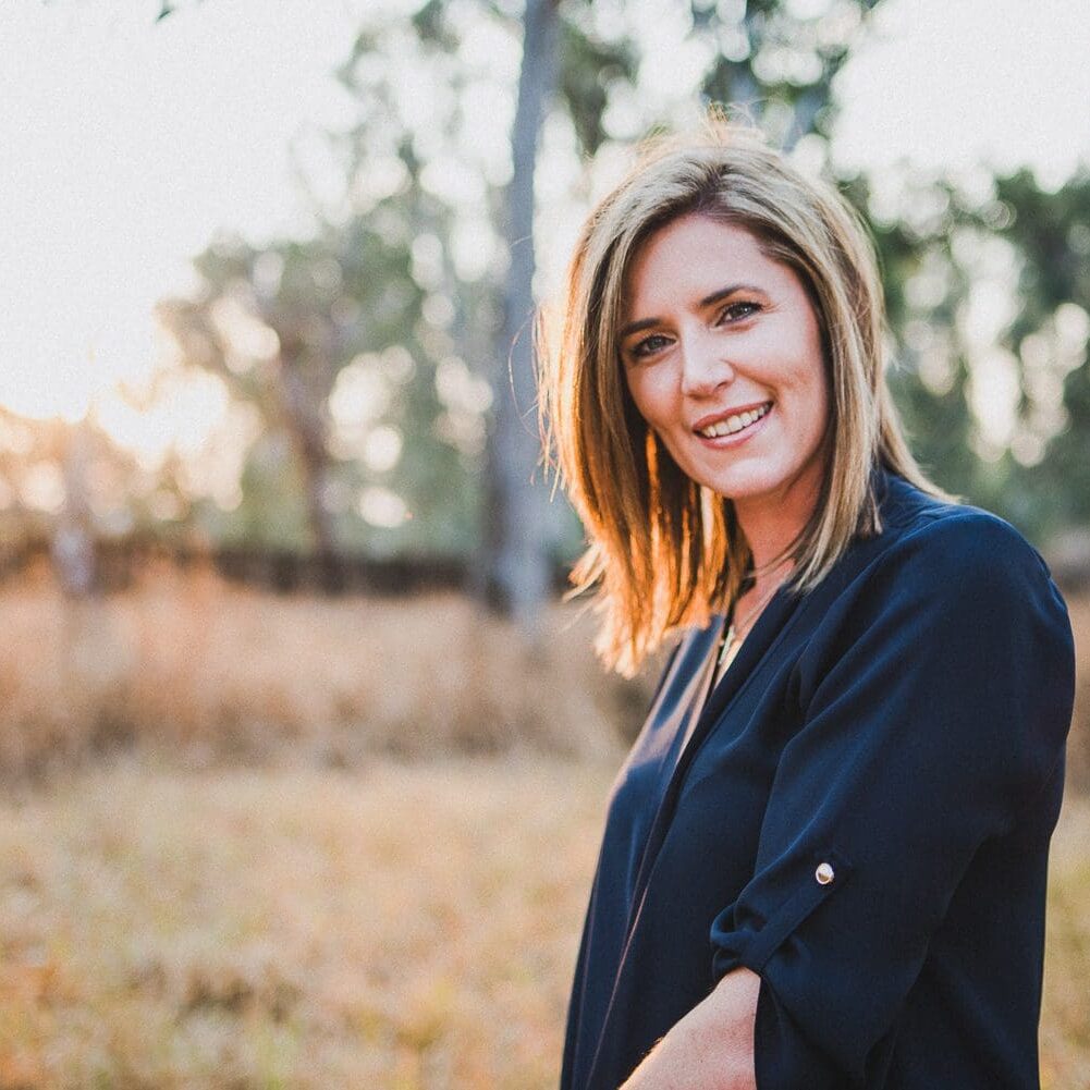 A woman with shoulder-length hair smiling outdoors in a field during sunset.